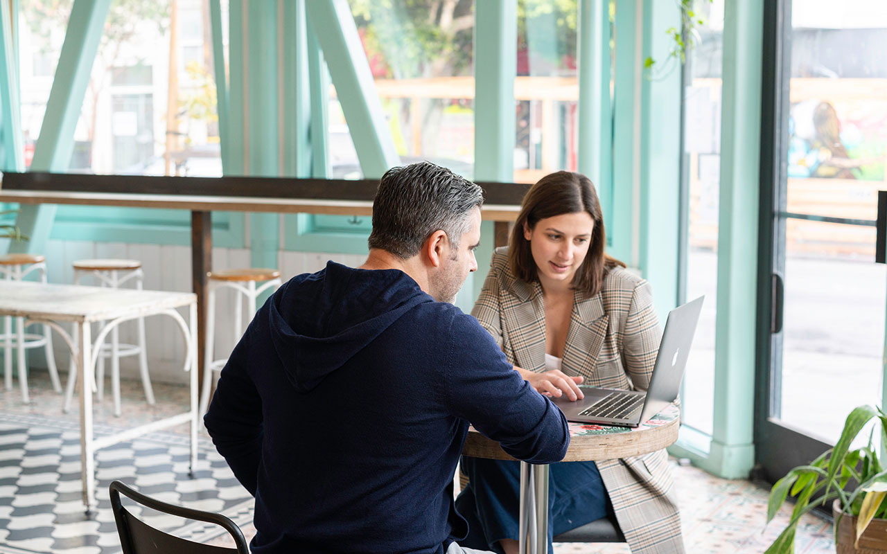Two professionals discussing over a laptop at a cafe with a bright, airy ambiance.